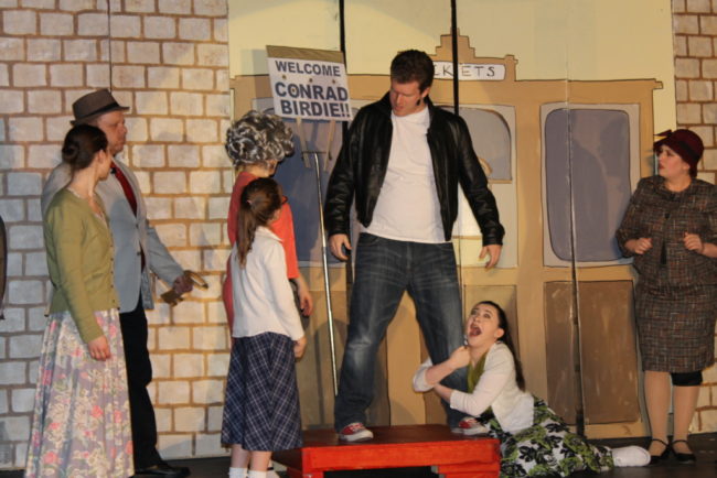 Matt Peterson (center) as Conrad Birdie with Olivia Winter as Ursula Merkel clinging to his leg with the Sweet Apple teens and parents