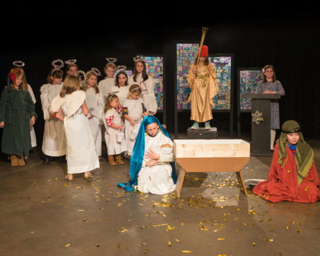 Lottie Doughty (kneeling left of the manger) as Imogene Herdman with Alice Levings (standing center above the manger) as Gladys Herdman, and Noah Davenport (kneeling right of the manger) as Ralph Herdman and the cast of The Best Christmas Pageant Ever at Silver Spring Stage