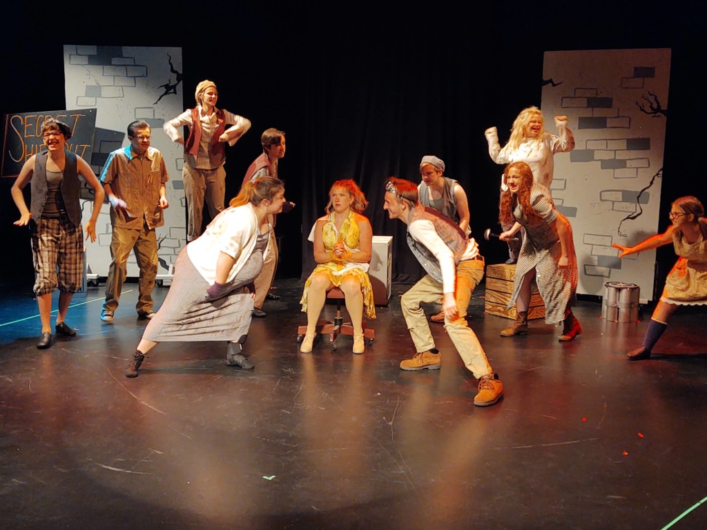 (Center- L to R) Kate Lawrence as Little Becky Two-Shoes, Eleanor Parks as Hope Cladwell, and Robert Dunham as Hot Blades Harry with the ensemble of Urinetown.