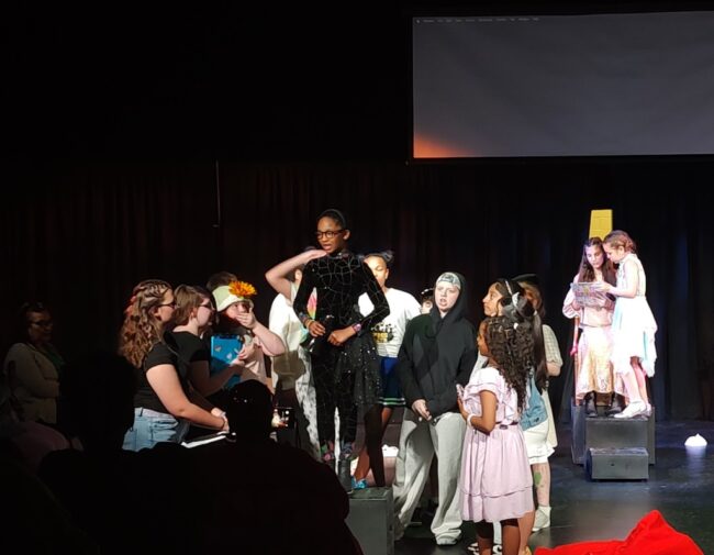 Greyson Herbert (standing center) as Miss Cue and the cast of Live Theatre at Apex Arts
