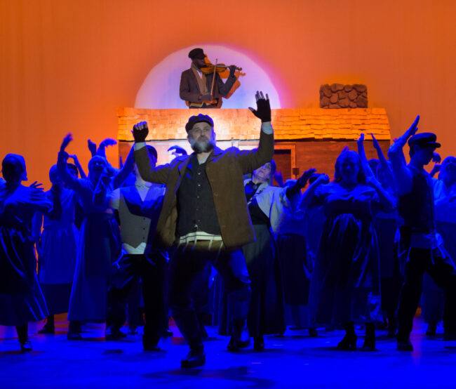 John Bruns (center) as Tevye and Michael Gabdamoshi (above) as The Fiddler with the ensemble of Fiddler on the Roof at HCST đˇ Neil Rubino